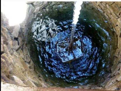 Метална кофа на верига, спусната в кладенец с вода; стените на шахтата са от камък.
